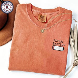 May include: A coral-colored t-shirt with the words "SOCIAL BATTERY" and a low battery graphic embroidered on the front. A gold necklace with a pendant rests on the shirt. The shirt has a tag that reads "COMFORT COLORS".