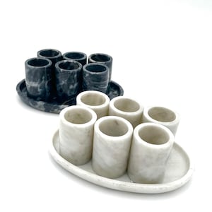 May include: Two sets of marble shot glasses on oval trays. One set is a dark gray with white veining, and the other is white with gray veining. Each set includes six shot glasses. The shot glasses are cylindrical.