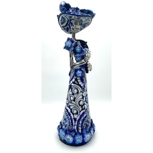May include: A blue and white Day of the Dead figurine of a female skeleton wearing a hat. The figurine has a detailed floral pattern on the dress and hat. The skeleton is holding a bouquet of flowers.