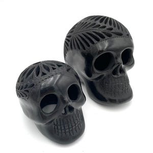May include: Two black decorative skull sculptures. The skulls have carved floral patterns on the top and around the eye sockets. The skulls are made of a glossy, dark material and are set against a white background.