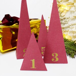 Może przedstawiać: Cztery czerwone papierowe ozdoby choinkowe z złotymi cyframi 1, 3 i 5. Ozdoby są ułożone w kształcie piramidy na białym tle.