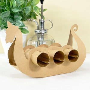 May include: A brown cardboard Viking ship toilet roll holder with three rolls of toilet paper. The ship features a detailed head and tail design. A green plant in a white pot and a glass water sprayer are in the background.