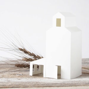 May include: A white paper model of a silo with a small window and a door. The model is sitting on a wooden surface with dried wheat stalks in the foreground.
