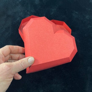Pu&ograve; includere: Una scatola di carta rossa a forma di cuore con un coperchio piegato. La scatola &egrave; fatta di carta spessa e ha una superficie testurizzata.