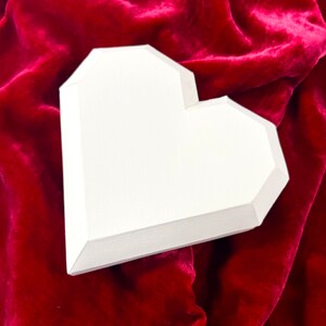 Pu&ograve; includere: Una scatola bianca a forma di cuore in cartone si trova su una superficie di velluto rosso. La scatola &egrave; vuota e ha una finitura liscia e opaca.