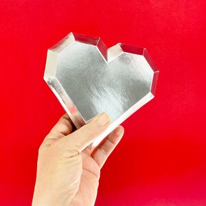 Pu&ograve; includere: Un vassoio a forma di cuore argentato con un design geometrico. Il vassoio &egrave; realizzato in un materiale riflettente e ha una superficie liscia.