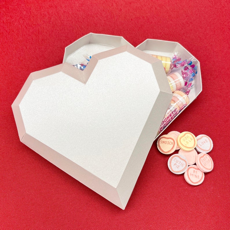Pu&ograve; includere: Una scatola bianca a forma di cuore con il coperchio aperto, che rivela una variet&agrave; di cuori di caramelle colorati all'interno. I cuori di caramelle hanno messaggi come "Smooch", "Cutie Pie" e "Be Mine".