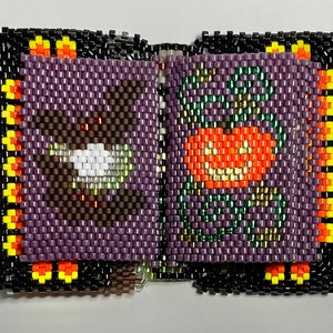 Op de afbeelding: Een klein kralenboek met een paars, groen, oranje en geel ontwerp. Het boek is gemaakt van kleine kralen die aan elkaar zijn genaaid om een ​​gedetailleerd patroon te creëren.