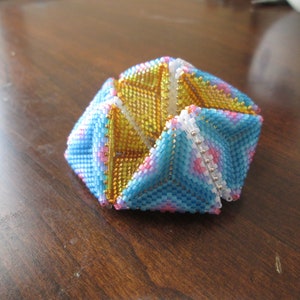 Könnte beinhalten: Ein vielseitiger, geometrischer Perlen-Schmuck mit einem Farbschema in Blau, Rosa, Weiß und Gold. Das Ornament besteht aus kleinen Perlen, die in einem komplexen Muster miteinander verwoben sind.