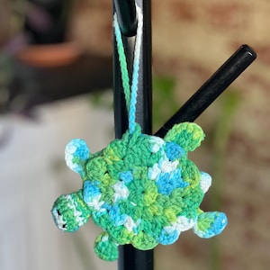 May include: A handcrafted crochet turtle decoration in green, blue, and white. The turtle features a textured shell and small flippers. It is suspended by a white and green string, ideal for hanging as an ornament.