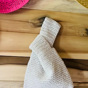 May include: A cream-colored, crocheted handbag with a knotted handle. The bag is made of a textured, open-weave fabric. The background includes a wooden surface and the edge of a pink and yellow straw hat.