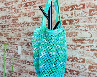 May include: A handmade, open-weave crochet tote bag in shades of blue, green, and white hangs from a black coat rack. The bag has a round bottom and two long handles. The background is a brick wall.