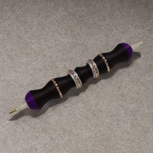 Könnte beinhalten: Ein schwarzer Diamant-Malstift mit einem lila Ende und einer weißen Spitze. Der Stift hat drei dekorative Ringe mit goldfarbenen Strasssteinen und weißen Akzenten. Der Stift ist für Präzision und Detailarbeit konzipiert.