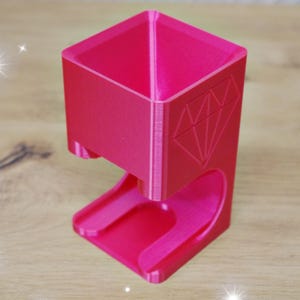 Può includere: Un organizer da scrivania rosa acceso, stampato in 3D. L'organizer ha una parte superiore quadrata, un design a forma di diamante sul lato e una base curva. L'articolo è in plastica e poggia su una superficie di legno.