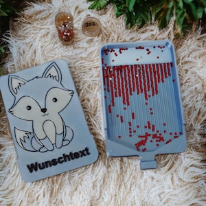 Puede incluir: Un kit de manualidades azul claro con un diseño de zorro de dibujos animados y el texto "Wunschtext". El kit incluye una bandeja llena de cuentas rojas, dos pequeños recipientes con cuentas y una superficie blanca y esponjosa.