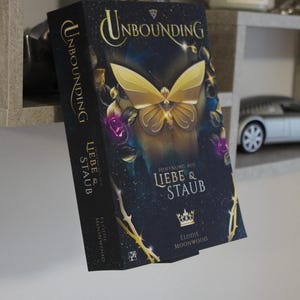 Op de afbeelding: Een hardcover boek getiteld "Unbounding" met een gouden vlinderillustratie op de omslag. Het boek heeft een donkerblauwe achtergrond met de tekst "Hoffnung aus Liebe & Staub" en de naam van de auteur, Elodie Moonwood.