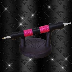 Könnte beinhalten: Ein schwarzer und pinkfarbener Diamantmalstift mit passendem Ständer. Der Stift hat einen schwarzen Griff mit pinkfarbenen Akzenten und eine weiße Spitze. Der Ständer ist schwarz und hat ein Diamantmuster.