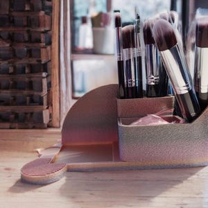 Puede incluir: Un soporte para brochas de maquillaje con un esquema de color degradado, que contiene varias brochas de maquillaje. El soporte es una mezcla de rosa, morado y dorado. Las brochas tienen mangos negros y cerdas suaves de color marrón. El soporte está sobre una superficie de madera.