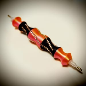 Puede incluir: Un bolígrafo decorativo para pintar con diamantes con un cuerpo degradado rosa y naranja, acentuado con bandas negras y pedrería. El bolígrafo tiene una punta transparente y una punta metálica dorada.