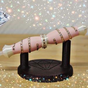 Könnte beinhalten: Ein rosa Diamant-Malstift mit Strass-Akzenten, präsentiert auf einem schwarzen Ständer. Der Stift ist mit mehreren Reihen von Strasssteinen und einer transparenten Spitze verziert. Eine Diamantgrafik befindet sich im Hintergrund.