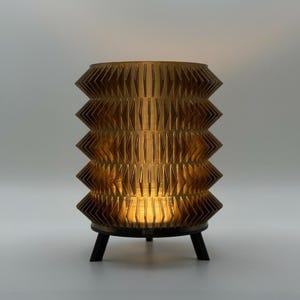 Puede incluir: Una lámpara de mesa decorativa con un diseño geométrico único. La lámpara tiene forma cilíndrica con una serie de bordes puntiagudos en capas. La lámpara es de color dorado cálido y se asienta sobre una base negra con tres patas. La lámpara está iluminada desde dentro, emitiendo un brillo cálido.