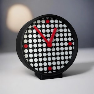 May include: A black and white dot pattern clock with red hands and accents. The clock has a round face and a square base. The clock's hands are red, and the dots on the face are white. The clock is a modern design.