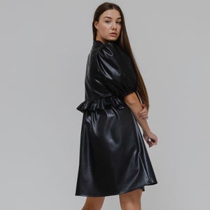 以下が含まれることがあります： 短いパフスリーブとウエストにフリルがあしらわれた黒のフェイクレザーのドレス。黒のコンバットブーツと合わせています。モデルは無地の白い背景の前に立っています。