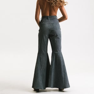 Könnte beinhalten: Eine graue Schlaghose. Die Hose hat eine hohe Taille und einen weiten Schlagstil. Das Model ist von hinten zu sehen, mit langen, welligen blonden Haaren. Der Hintergrund ist schlicht weiß.