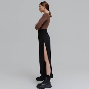 Pode incluir: Uma mulher vestindo uma blusa marrom de manga comprida e calças pretas com uma fenda alta. As calças são combinadas com botas pretas com solas grossas. A roupa é simples e moderna, com foco em linhas limpas e uma estética minimalista.