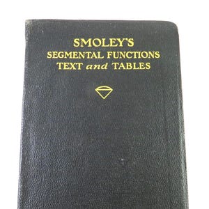 May include: A vintage black book titled "Smoley's Segmental Functions Text and Tables" with gold lettering. The cover features a diamond symbol and the text "First Edition". The book has a worn appearance.