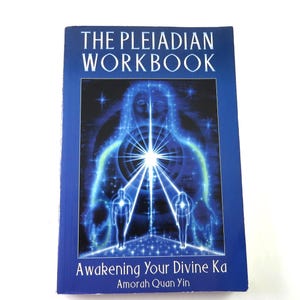 May include: A blue book titled "THE PLEIADIAN WORKBOOK" with the subtitle "Awakening Your Divine Ka" by Amorah Quan Yin. The cover features a glowing blue illustration of a figure with radiating light and two smaller figures.