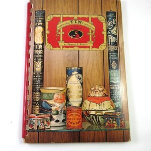 May include: A vintage book titled "Collectible Tin Containers with Price Guide" by Ernest L. Pettit. The cover features a red and gold design. The image also shows various antique tin containers, including a smiling figure and a cylindrical container.
