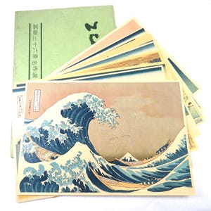 May include: A collection of Japanese art prints featuring a famous wave design with boats, a mountain, and a green booklet with Japanese text. The prints have a vintage aesthetic with blue, beige, and pink hues.
