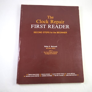 May include: A maroon softcover book titled The Clock Repair FIRST READER: SECOND STEPS for the BEGINNER by Philip E. Balcomb. The cover features yellow and white text listing topics including bench practices, tools, and clock restoration.
