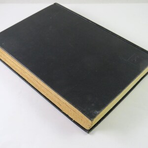 Może przedstawiać: Zamknięta książka w twardej oprawie z czarną okładką i poż&oacute;łkłymi, postarzanymi stronami. Grzbiet książki jest widoczny, a na okładce znajduje się mały, wytłoczony wz&oacute;r. Książka leży na białej powierzchni.