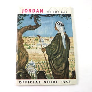 May include: Vintage 1956 Official Guide to Jordan. The cover features an illustration of a person in traditional attire overlooking a city with domes and trees. The title reads "Jordan The Holy Land".