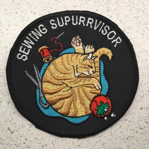 May include: A black and white embroidered patch with a cat sleeping on a blue background. The text "Sewing Supurrvvisor" is written in a white arc around the cat.