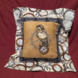 May include: A decorative pillow with a brown and beige fabric featuring a white owl with a top hat and a clock face. The pillow is surrounded by a border of clocks and gears.