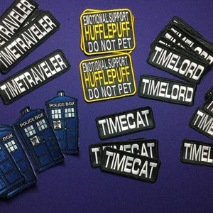 Könnte beinhalten: Eine Sammlung von Bügelbildern mit verschiedenen Designs, darunter ein blauer TARDIS, ein Time Lord, eine Time Lady, ein Time Traveler und eine Time Cat. Die Patches sind schwarz, weiß und gelb mit weißem Text. Die Patches befinden sich auf einem violetten Hintergrund.