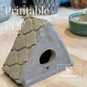 Puede incluir: Una casita para pájaros en miniatura gris hecha a mano, con un tejado texturizado y un agujero de entrada circular. La casita para pájaros es de forma triangular y tiene las palabras "Printable PDF Miniature Birdhouse" en el fondo.