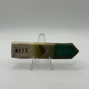 May include: A ceramic arrow-shaped object with a textured surface. The arrow is divided into three sections: a light beige section with the numbers "d111", a brown and yellow section, and a dark green arrowhead. The object is displayed on a clear plastic stand.