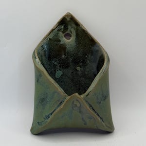 May include: A wall-mounted ceramic pocket planter with a pointed top and a circular hole for hanging. The exterior features a matte sage green finish, while the interior is glazed in a deep, glossy, mottled dark green and black speckled texture.
