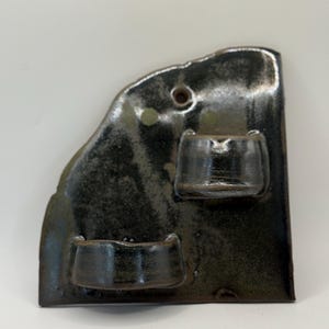 May include: A wall-mounted ceramic planter with a dark, glossy, mottled black and brown glaze. The piece features an irregular, asymmetrical shape with two integrated semi-circular pockets for holding small items or plants and a single hole at the top.