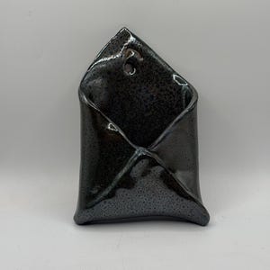 Puede incluir: Un bolsillo de cerámica para pared con un esmalte brillante de color gris carbón oscuro y textura moteada. El objeto tiene forma de sobre con una solapa superior puntiaguda y un pequeño orificio circular cerca del vértice para colgarlo.