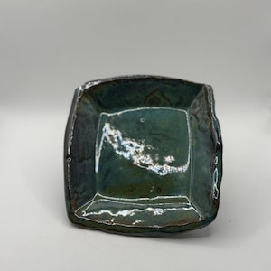 May include: A handmade, square ceramic dish with a dark teal glaze. The dish has a slightly irregular shape with textured edges and a glossy finish. The interior has a lighter, almost iridescent quality.