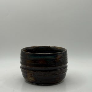 May include: A handcrafted, dark brown ceramic bowl with horizontal textured lines. The bowl has a rounded shape and a dark interior, with hints of green and brown. The exterior has a rustic, earthy appearance.