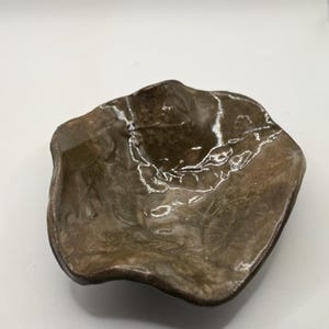 Op de afbeelding: Een handgemaakte keramische kom met een organische vorm en een glanzend, gevlekt bruin en grijs glazuur. De kom heeft een onregelmatige rand en een ondiep, hol interieur, perfect voor het serveren van kleine porties of als decoratief stuk.