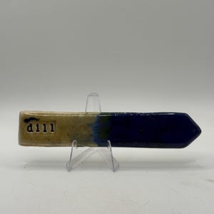 May include: A ceramic garden marker with a rectangular top and an arrow-shaped end. The top is tan with the word "dill" and the number "111" printed in black. The marker transitions to a dark blue colour. It is displayed on a clear plastic stand.