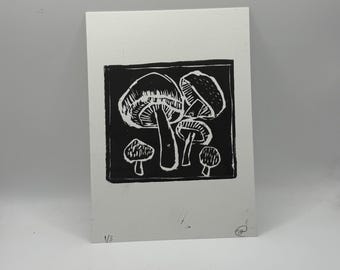 Puede incluir: Un grabado en linóleo en blanco y negro que representa un grupo de setas. La impresión muestra varias formas y tamaños de setas, con texturas detalladas y tonos contrastantes. La obra de arte está sobre un papel rectangular blanco.