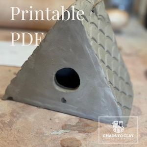 May include: A grey ceramic birdhouse with a triangular roof and a round entry hole. The roof has a textured, scale-like pattern. The text "Printable PDF" is visible on the front. A twine loop is attached for hanging.